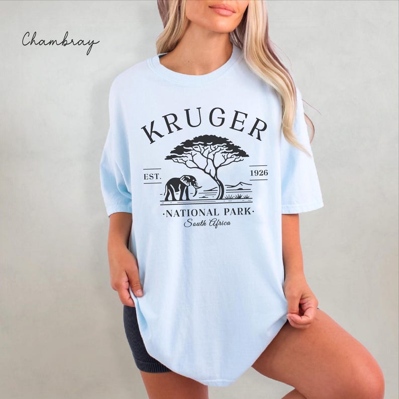Puede incluir: Camiseta azul claro con un gr&aacute;fico negro de un elefante, un &aacute;rbol y las palabras "KRUGER NATIONAL PARK South Africa". La camiseta tambi&eacute;n tiene el texto "EST. 1926" encima y debajo del gr&aacute;fico. La palabra "Chambray" est&aacute; escrita en cursiva en la esquina superior izquierda.