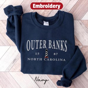 Pode incluir: Camisola azul marinho com bordado "Outer Banks, Carolina do Norte" e um gr&aacute;fico de farol. A camisola tamb&eacute;m apresenta os n&uacute;meros "15" e "87". A palavra "Embroidery" est&aacute; num banner vermelho no topo.