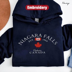 May include: Navy blue hooded sweatshirt with white embroidered text reading "Niagara Falls", a red maple leaf, and "Ontario Canada". The word "Embroidery" is printed in red. The sweatshirt is a Gildan brand.