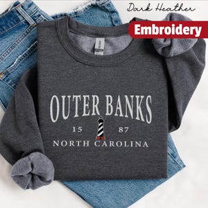 Pode incluir: Camisola cinza escuro com o texto branco "OUTER BANKS", um desenho de farol e "NORTH CAROLINA". Uma faixa vermelha diz "Embroidery". A camisola est&aacute; dobrada sobre uns jeans azuis.