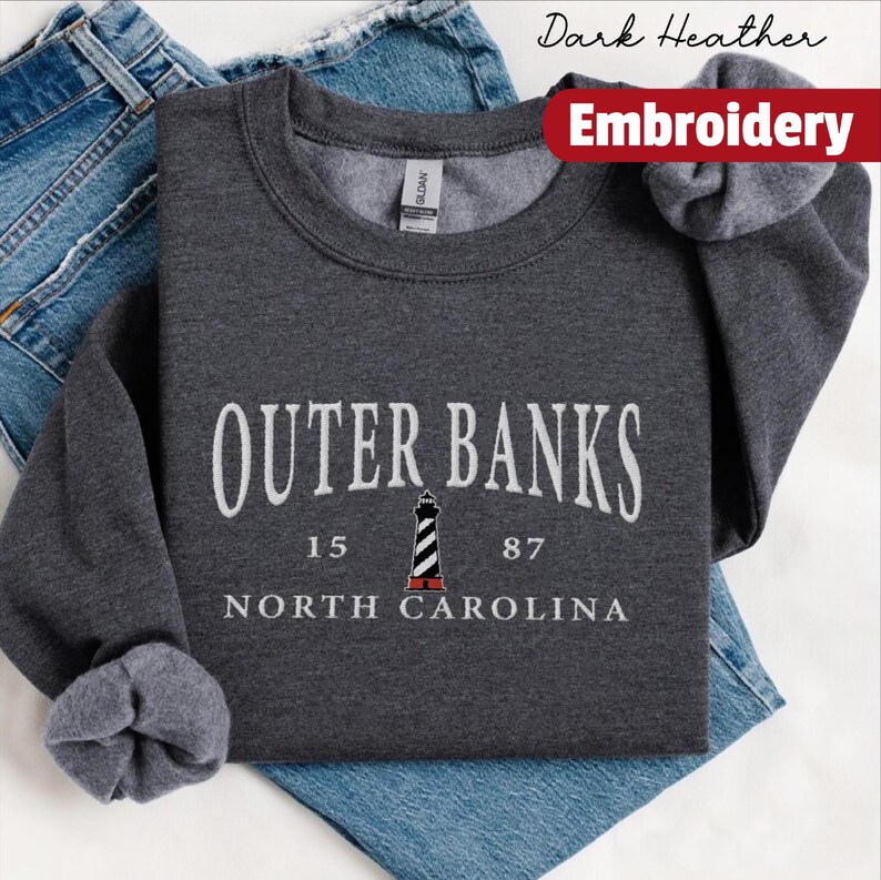 Pode incluir: Camisola cinza escuro com o texto branco "OUTER BANKS", um desenho de farol e "NORTH CAROLINA". Uma faixa vermelha diz "Embroidery". A camisola est&aacute; dobrada sobre uns jeans azuis.