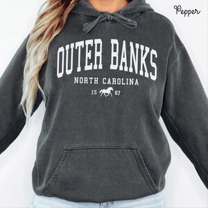 May include: Dark gray Outer Banks North Carolina hoodie with white lettering. The hoodie features a drawstring hood, a kangaroo pocket, and the text "Outer Banks North Carolina" with a horse graphic and the numbers "15 87".