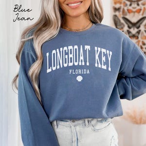 May include: A blue jean colored sweatshirt with the words "LONGBOAT KEY FLORIDA" in white block letters. A seashell graphic is below the word "FLORIDA". The words "Blue Jean" are written in cursive in the upper left corner.