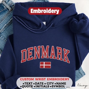 May include: A navy blue hooded sweatshirt featuring the word DENMARK embroidered in red with a white outline, positioned above a small red and white Danish flag. The text Embroidery appears at the top, and custom wrist embroidery options are listed.