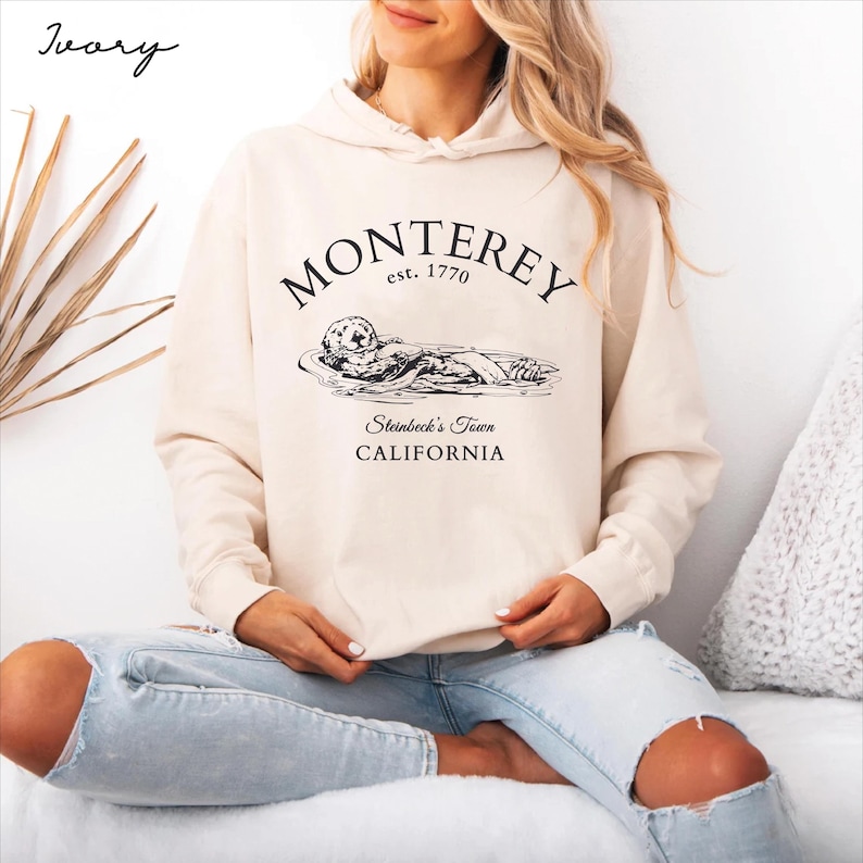 May include: A cream-coloured hooded sweatshirt with the text "Monterey est. 1770 Steinbeck's Town CALIFORNIA" and a black and white illustration of a sea otter.