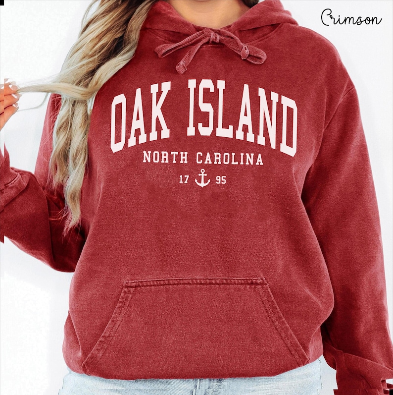 May include: Crimson red hoodie with a drawstring hood and a front pocket. The white text on the front reads "OAK ISLAND NORTH CAROLINA 17 ⚓ 95". The hoodie is a solid colour and has a relaxed fit.