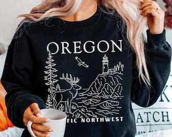Può includere: Una felpa nera a maniche lunghe con una grafica a linee bianche che raffigura un cervo, un faro su una scogliera, un uccello in volo e alberi di pino. Il testo OREGON e PACIFIC NORTHWEST è stampato in bianco sulla parte del petto.