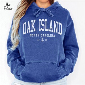 May include: A blue hooded sweatshirt with the text "OAK ISLAND NORTH CAROLINA" in white block letters. Below the text is "17 95" with an anchor symbol. The sweatshirt has a front pocket and a drawstring hood.