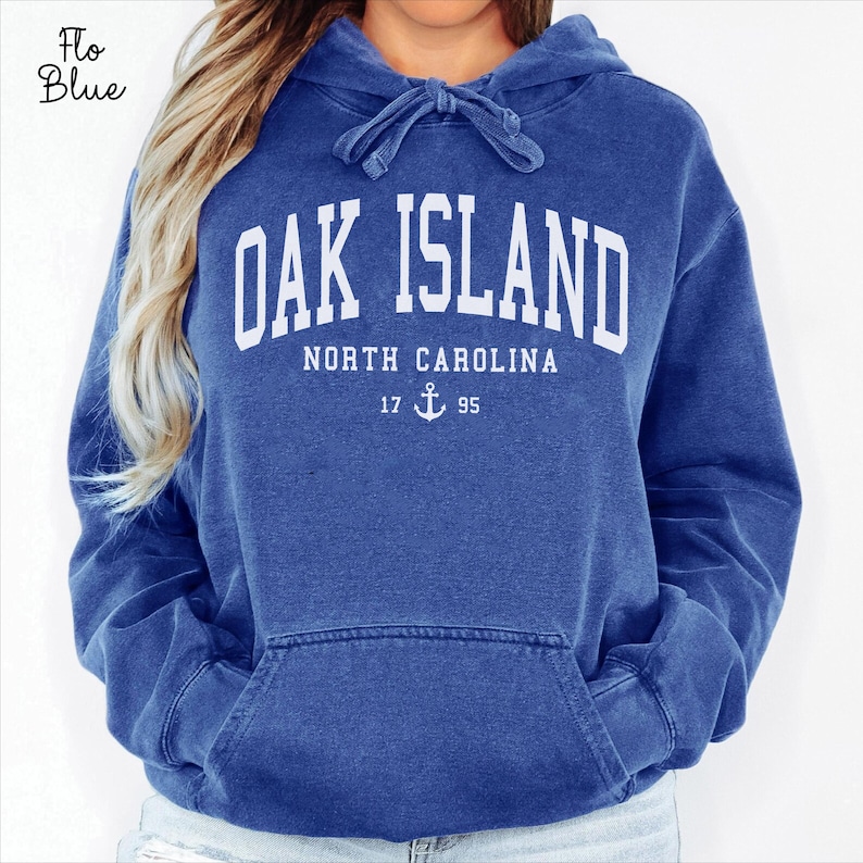 May include: A blue hooded sweatshirt with the text "OAK ISLAND NORTH CAROLINA" in white block letters. Below the text is "17 95" with an anchor symbol. The sweatshirt has a front pocket and a drawstring hood.