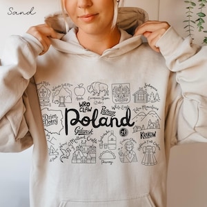 May include: Sand-colored hoodie with a black and white graphic of Poland's landmarks and the word "Poland" in a bold font. The hoodie features a drawstring hood and a kangaroo pocket.