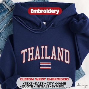 May include: A navy blue hooded sweatshirt featuring the word THAILAND embroidered in white with red outlines in an arched font. Below the text is an embroidered flag of Thailand. The hoodie includes a drawstring hood and long sleeves.