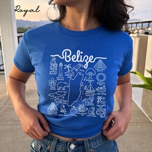 Pode incluir: Uma t-shirt de manga curta azul royal com uma impressão gráfica branca de um mapa do Belize rodeado por ilustrações de marcos locais, vida selvagem e atividades, incluindo etiquetas de texto para cada ícone representado.