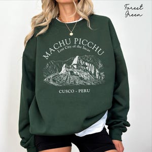 Peut inclure: Un sweat-shirt à col rond vert forêt avec une impression graphique blanche représentant les ruines du Machu Picchu. Le texte indique "MACHU PICCHU Lost City of the Incas" et "CUSCO - PERU". Le motif inclut un dessin au trait du paysage.