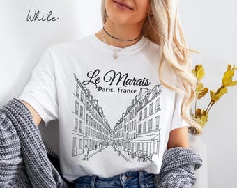 May include: White t-shirt featuring a black and white line drawing of a street scene in Paris, France. The text "Le Marais, Paris, France" is above the illustration. The word "White" is written in cursive in the upper left corner.