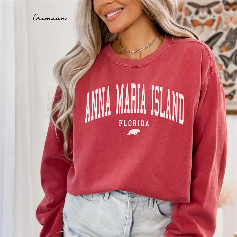 May include: Crimson red sweatshirt with the text "ANNA MARIA ISLAND FLORIDA" in white block letters. A small white sea turtle graphic is below the text. The sweatshirt is a crew neck style.