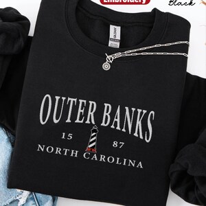 Pode incluir: Camisola preta com texto bordado branco que diz "Outer Banks, Carolina do Norte" e um gr&aacute;fico de farol. Um colar de prata com um pingente circular repousa sobre a camisa. A palavra "Bordado" est&aacute; em um c&iacute;rculo vermelho.