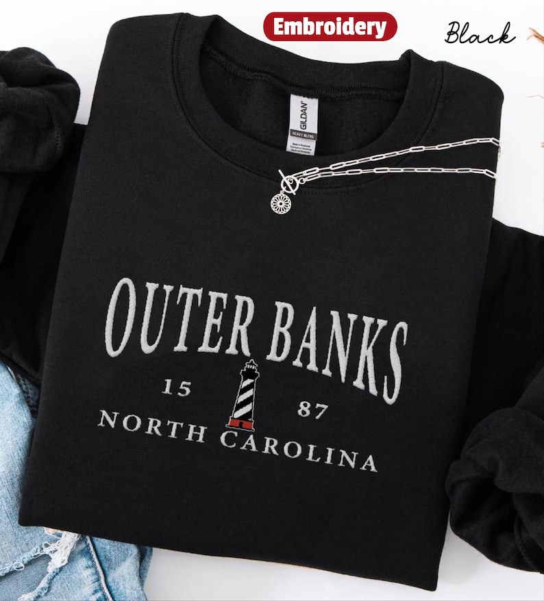 Pode incluir: Camisola preta com texto bordado branco que diz "Outer Banks, Carolina do Norte" e um gr&aacute;fico de farol. Um colar de prata com um pingente circular repousa sobre a camisa. A palavra "Bordado" est&aacute; em um c&iacute;rculo vermelho.
