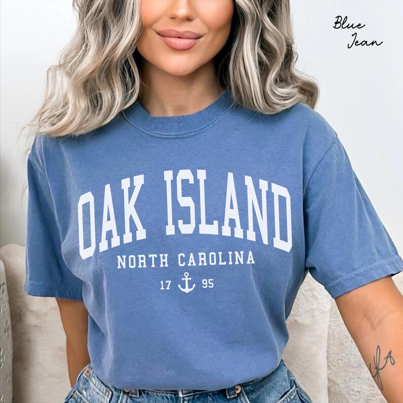 May include: A blue t-shirt featuring the text "OAK ISLAND" in white block letters. Underneath, it reads "NORTH CAROLINA" with the numbers "17 95" and an anchor symbol. The shirt is a solid blue colour.