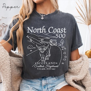 May include: Dark gray t-shirt with a white graphic of a road, a camper van, and a Highland cow. The text reads "North Coast 500" and "Scotland Scottish Highlands 516-mile (830 km)". The shirt is worn by a person.