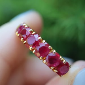 May include: A gold band ring featuring a row of five round-cut, deep red rubies. Each gemstone is securely held in place by small gold prongs, creating a continuous line of stones across the top of the ring, showcasing a vibrant and polished finish.