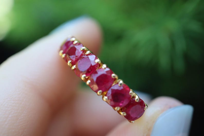 May include: A gold band ring featuring a row of five round-cut, deep red rubies. Each gemstone is securely held in place by small gold prongs, creating a continuous line of stones across the top of the ring, showcasing a vibrant and polished finish.