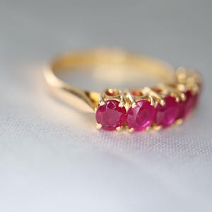 May include: A gold ring featuring a row of five round-cut, deep pinkish-red rubies set in individual prongs. The band is smooth and polished, showcasing a classic design that highlights the vibrant color and faceted texture of the gemstones.