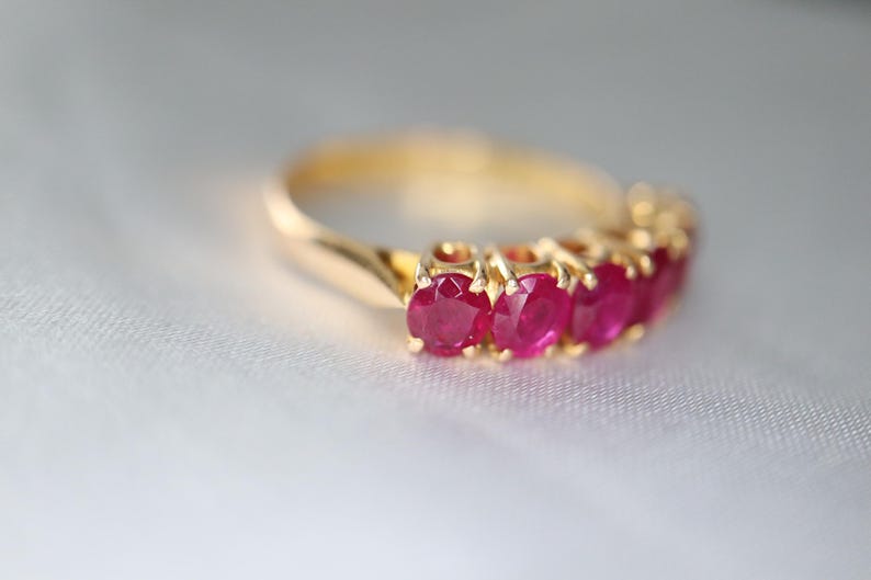 May include: A gold ring featuring a row of five round-cut, deep pinkish-red rubies set in individual prongs. The band is smooth and polished, showcasing a classic design that highlights the vibrant color and faceted texture of the gemstones.