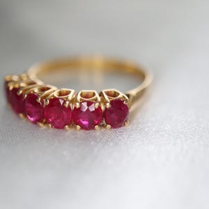 May include: A gold ring featuring five round, vibrant red rubies set in a row. The band is crafted from polished yellow gold, and each gemstone is held securely in place by individual metal prongs, creating a continuous, elegant line of stones.