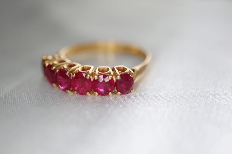 May include: A gold ring featuring five round, vibrant red rubies set in a row. The band is crafted from polished yellow gold, and each gemstone is held securely in place by individual metal prongs, creating a continuous, elegant line of stones.