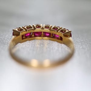 May include: A gold band ring featuring a row of five round, vibrant pinkish-red rubies set in individual metal prongs. The ring is captured from a side angle, highlighting the circular setting structure and the polished gold metal finish.