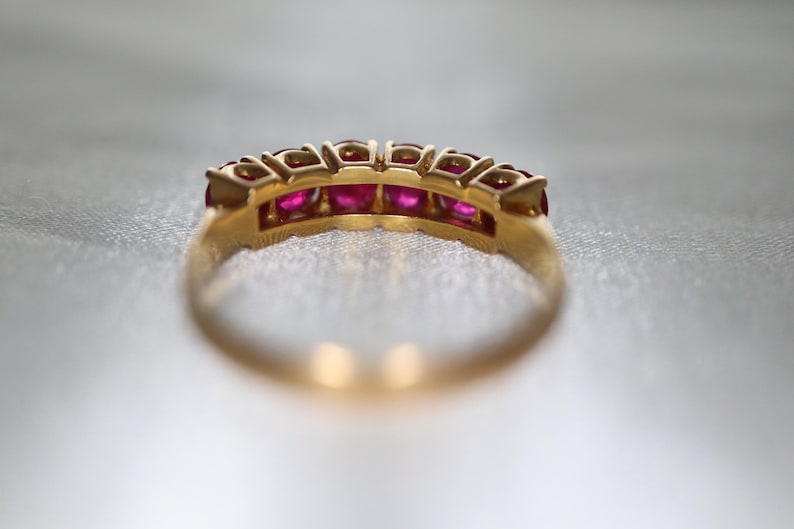 May include: A gold band ring featuring a row of five round, vibrant pinkish-red rubies set in individual metal prongs. The ring is captured from a side angle, highlighting the circular setting structure and the polished gold metal finish.