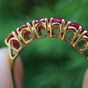 May include: A gold ring featuring five faceted, deep red rubies set in a row. The band is constructed with an open, arched gallery design that supports each stone individually, showcasing the metalwork and the vibrant color of the gemstones.