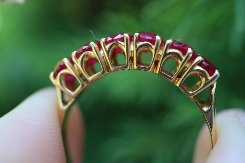 May include: A gold ring featuring five faceted, deep red rubies set in a row. The band is constructed with an open, arched gallery design that supports each stone individually, showcasing the metalwork and the vibrant color of the gemstones.