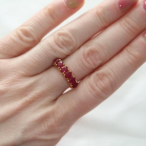 May include: A gold ring featuring oval-cut, vivid red gemstones. The ring is a band style, with the gemstones set closely together around the entire circumference. The ring is shown on a person's finger.
