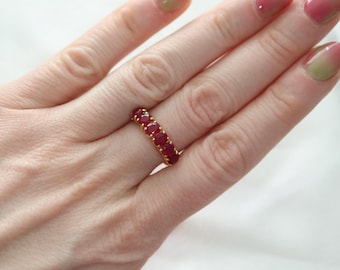 May include: A gold band ring featuring five round, deep red faceted gemstones set in a row. The ring is worn on a finger, showcasing the vibrant red stones and the polished gold metal setting that holds each individual gemstone securely in place.