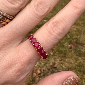 May include: A gold band ring featuring five round, deep red rubies set in a row. The stones are held in place by small gold prongs, creating a continuous line of vibrant red gems across the top of the band, showcasing a classic and elegant design.