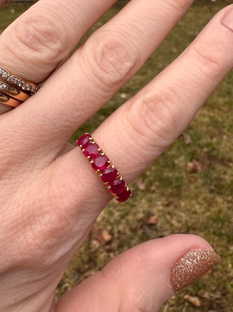 May include: A gold band ring featuring five round, deep red rubies set in a row. The stones are held in place by small gold prongs, creating a continuous line of vibrant red gems across the top of the band, showcasing a classic and elegant design.