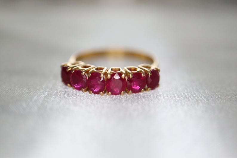 May include: A gold ring featuring a row of six oval-cut, deep pinkish-red rubies set in a prong setting. The band is crafted from polished yellow gold, showcasing a continuous line of faceted gemstones that catch the light across the surface.