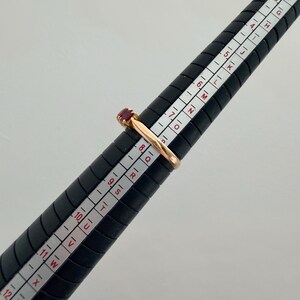 May include: A gold ring featuring a small, rectangular red gemstone set on a black ring mandrel. The ring is positioned at the size 8 mark on the mandrel, which displays both numerical and alphabetical sizing scales for accurate measurement.