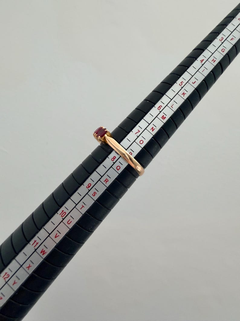 May include: A gold ring featuring a small, rectangular red gemstone set on a black ring mandrel. The ring is positioned at the size 8 mark on the mandrel, which displays both numerical and alphabetical sizing scales for accurate measurement.