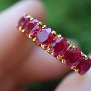 May include: A gold band ring featuring a row of six round-cut, deep red rubies set in individual prongs. The stones are arranged in a straight line along the top of the band, showcasing a faceted surface that reflects light across the red gems.