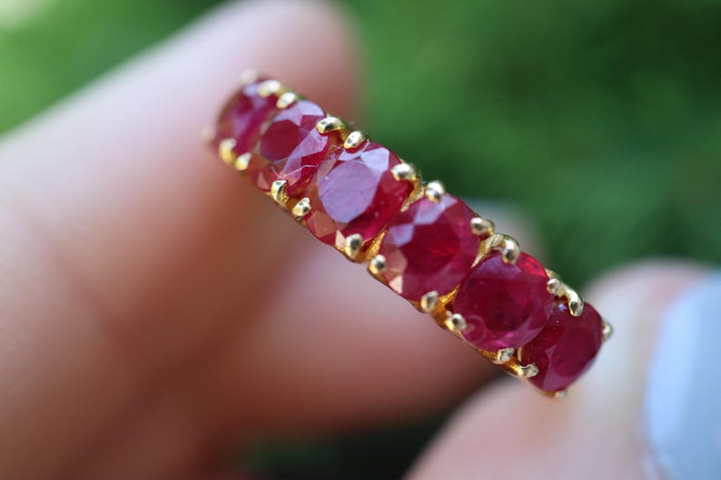 May include: A gold band ring featuring a row of six round-cut, deep red rubies set in individual prongs. The stones are arranged in a straight line along the top of the band, showcasing a faceted surface that reflects light across the red gems.