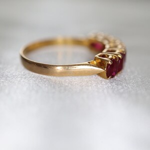 May include: A gold ring featuring a row of multiple oval-cut red rubies set in a prong setting. The band is smooth and polished, showcasing a classic design with the vibrant red gemstones aligned horizontally along the top of the metal band.