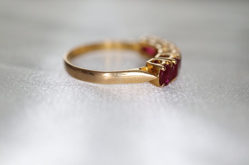 May include: A gold ring featuring a row of multiple oval-cut red rubies set in a prong setting. The band is smooth and polished, showcasing a classic design with the vibrant red gemstones aligned horizontally along the top of the metal band.