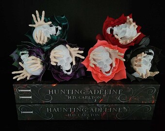 May include: A collection of decorative floral arrangements featuring white skulls with outstretched hands, set atop two books titled "Hunting Adeline" by H.D. Carlton. The flowers are in shades of green, purple, red, and black.