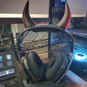 May include: Black gaming headset with devil horn attachments. The headset is resting on a black charging station. The horns are black with red tips. The headset has black earcups and a black headband.