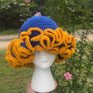 Könnte beinhalten: Eine gehäkelte Mütze mit einer blauen Krone und einem gelben, gekräuselten Rand. Die Mütze ist aus einem weichen, strukturierten Garn gefertigt und hat ein einzigartiges, verspieltes Design.