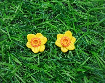May include: Two yellow flower-shaped earrings with orange centers, displayed on a green grassy surface.