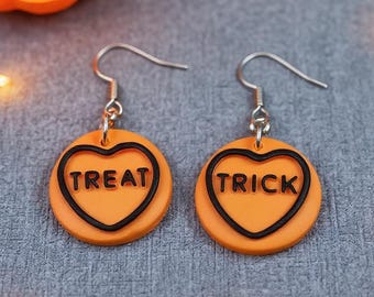 May include: A pair of orange circular drop earrings with silver-toned hooks. Each earring features a raised black heart outline containing the text TREAT on one earring and TRICK on the other, rendered in a bold, black sans-serif font.