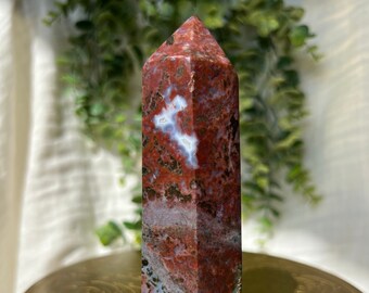 May include: A red, white, and brown agate crystal point, also known as a red jasper point, is displayed on a gold-colored table with intricate designs. The crystal is polished and has a pointed tip.